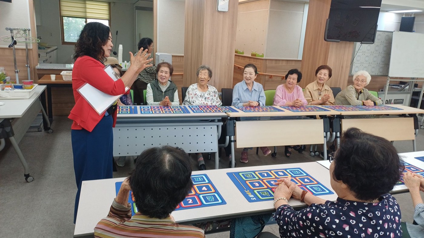 기관 수업에서 손 활동판을 앞에 두고 강사의 설명을 듣는 어르신들